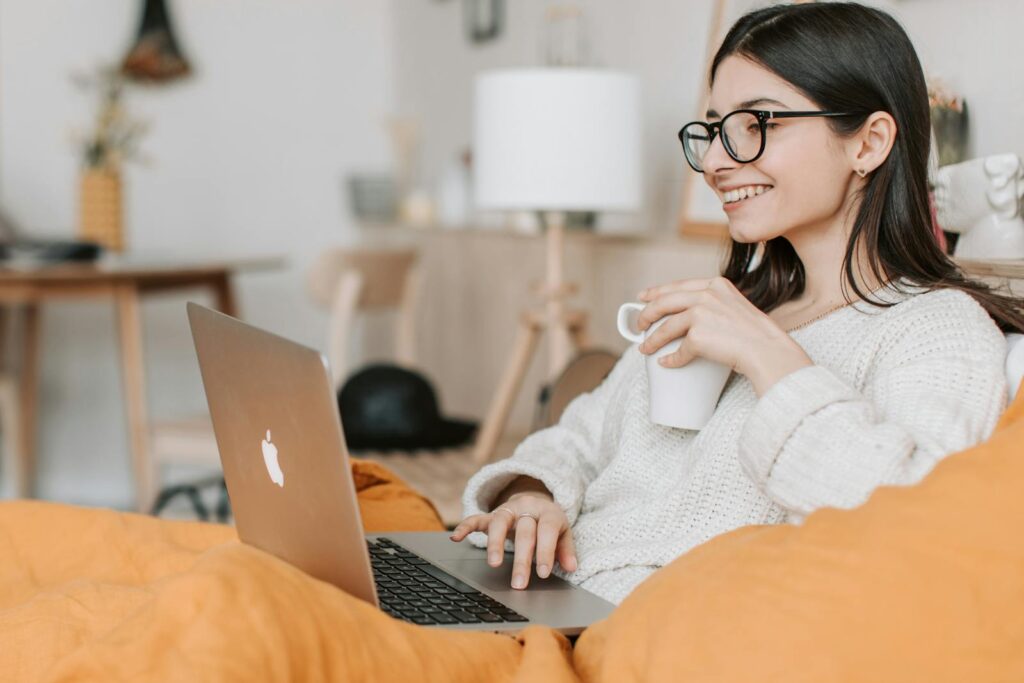 mulher branca cabelo comprido preto de óculos sentada ao sofá olhando o notebook e segurando uma xícara de café sorrindo para softwares para e-commerce