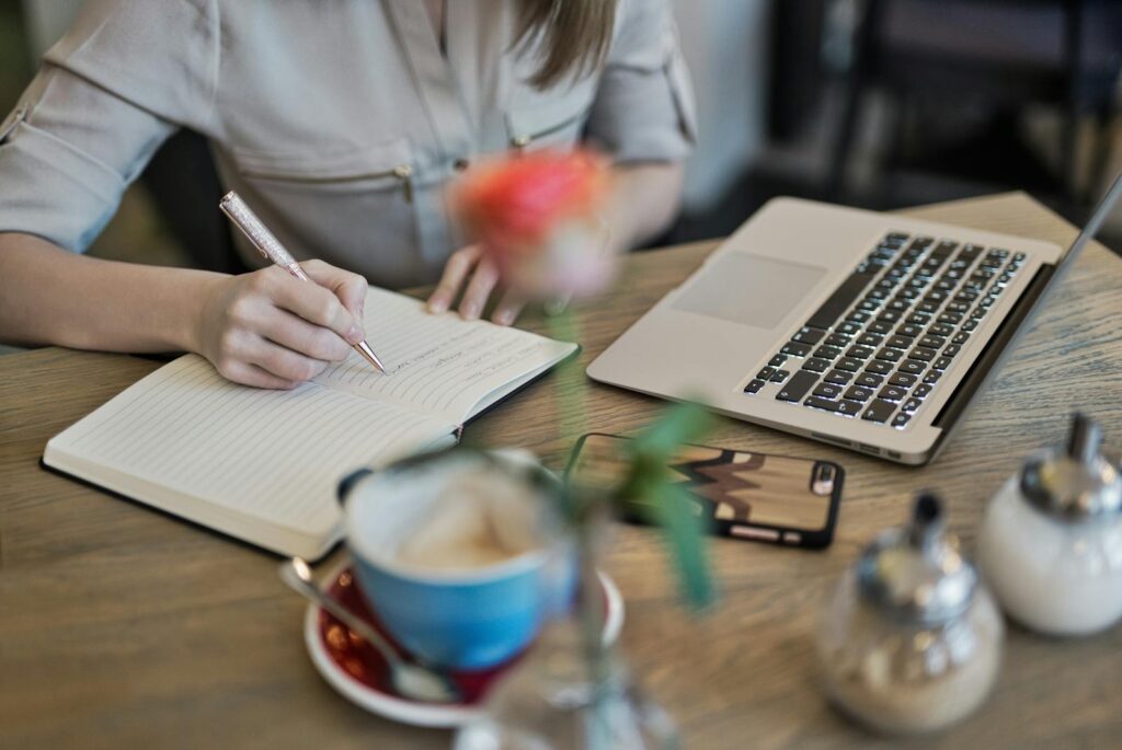 Pessoa senada na frente de uma mesa com notebook aberto. Na frente do notebook esta um caderno aberdo e a pessoa esta escrevendo alguma coisa nele. A mesa tem uma xícara azul em cima de um pires. A pessoa é profissional em UX Writing.