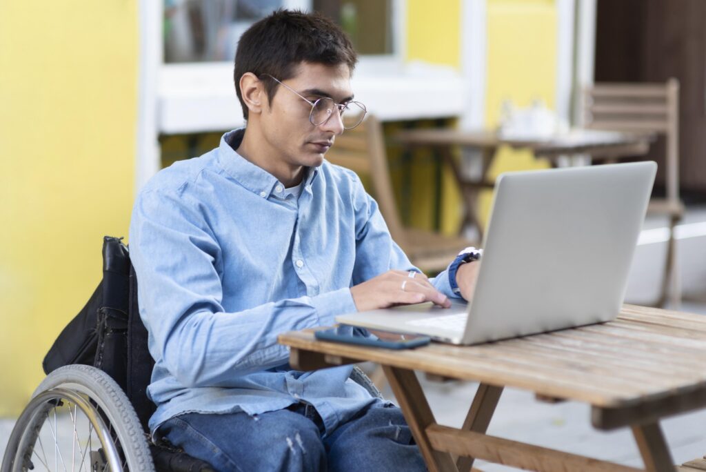 Homem cadeirante usando notebook em ambiente externo, representando o acesso a rotas acessíveis e recursos de acessibilidade digital como comandos por voz e mapas adaptados.